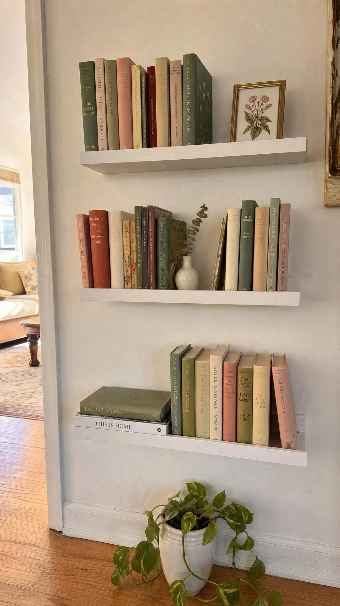 Three IKEA BEKVAM spice racks painted white and mounted as staggered floating bookshelves, styled with sage, blush, and terracotta book spines on a cozy white wall