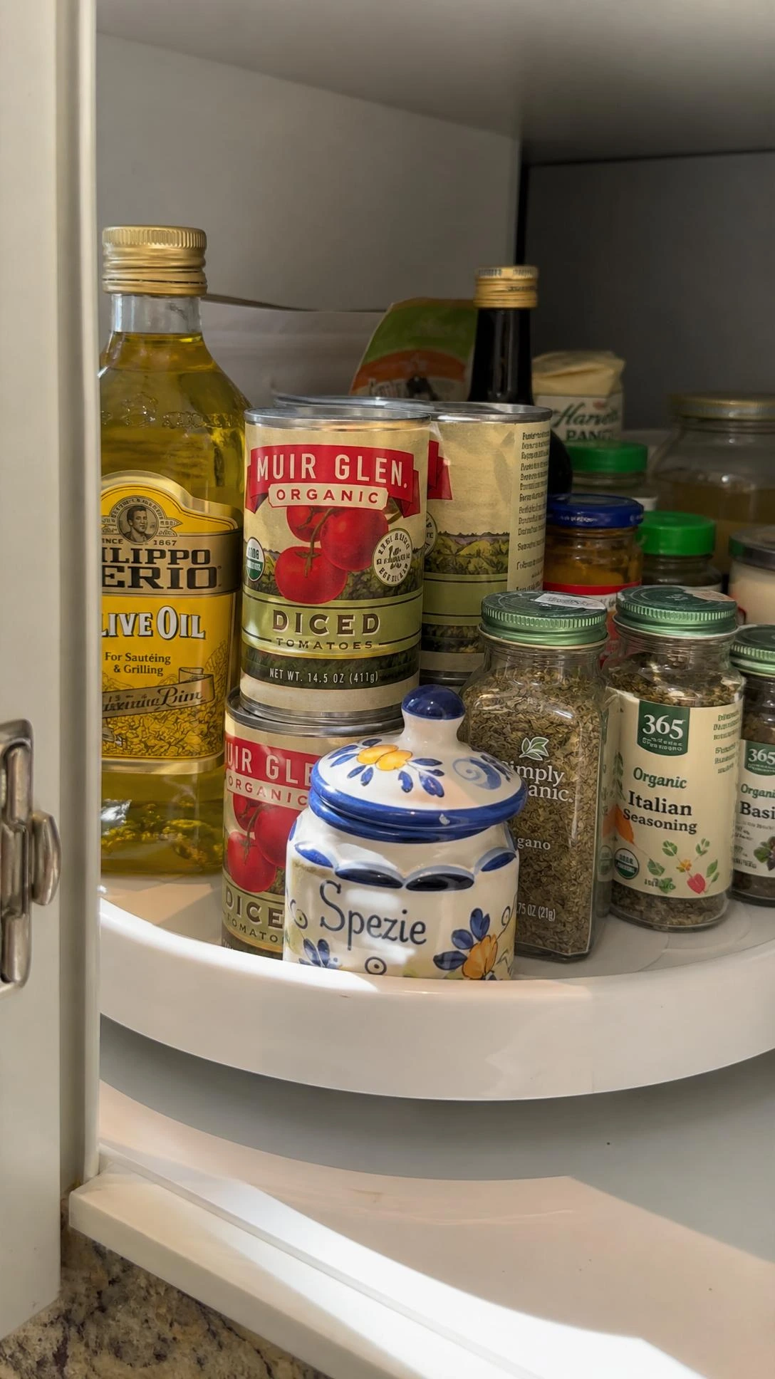 White Lazy Susan turntable inside a deep cabinet corner, stocked with tomato cans, olive oil, and spice jars — maximizing hard-to-reach kitchen storage