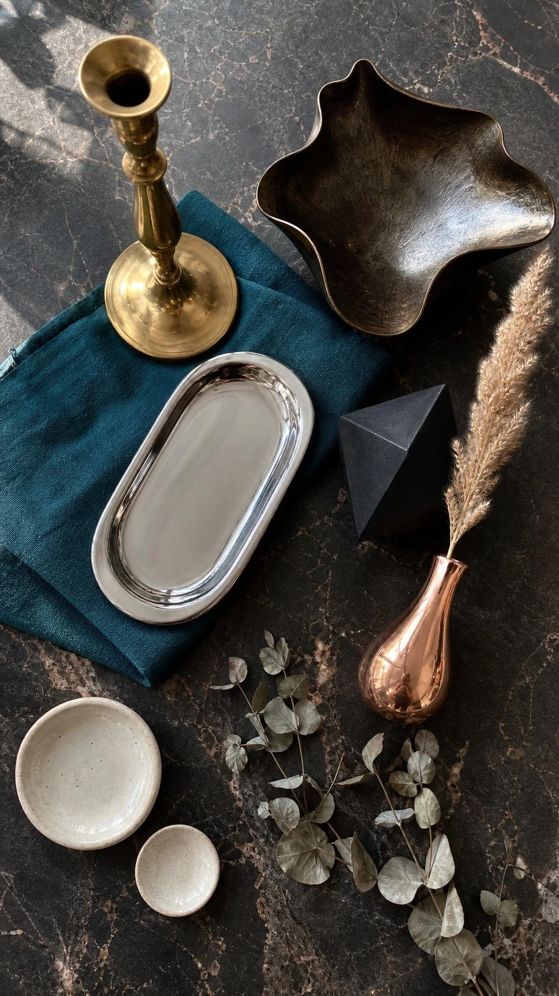 Styled flat lay of mixed metal decorative objects on dark marble — brushed brass, polished chrome, bronze, and matte black grouped with intentional contrast for an eclectic luxury look