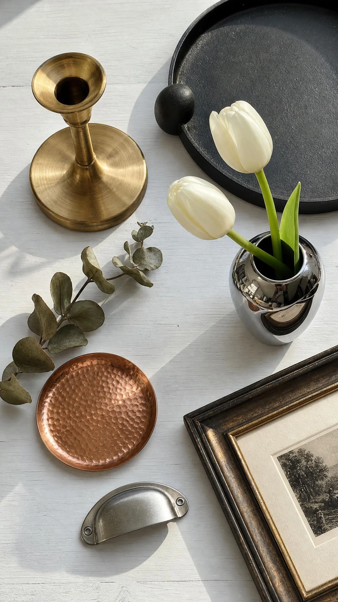 Flat lay of mixed metals on white wood surface — brushed brass candlestick, matte black tray, and polished silver objects styled together intentionally