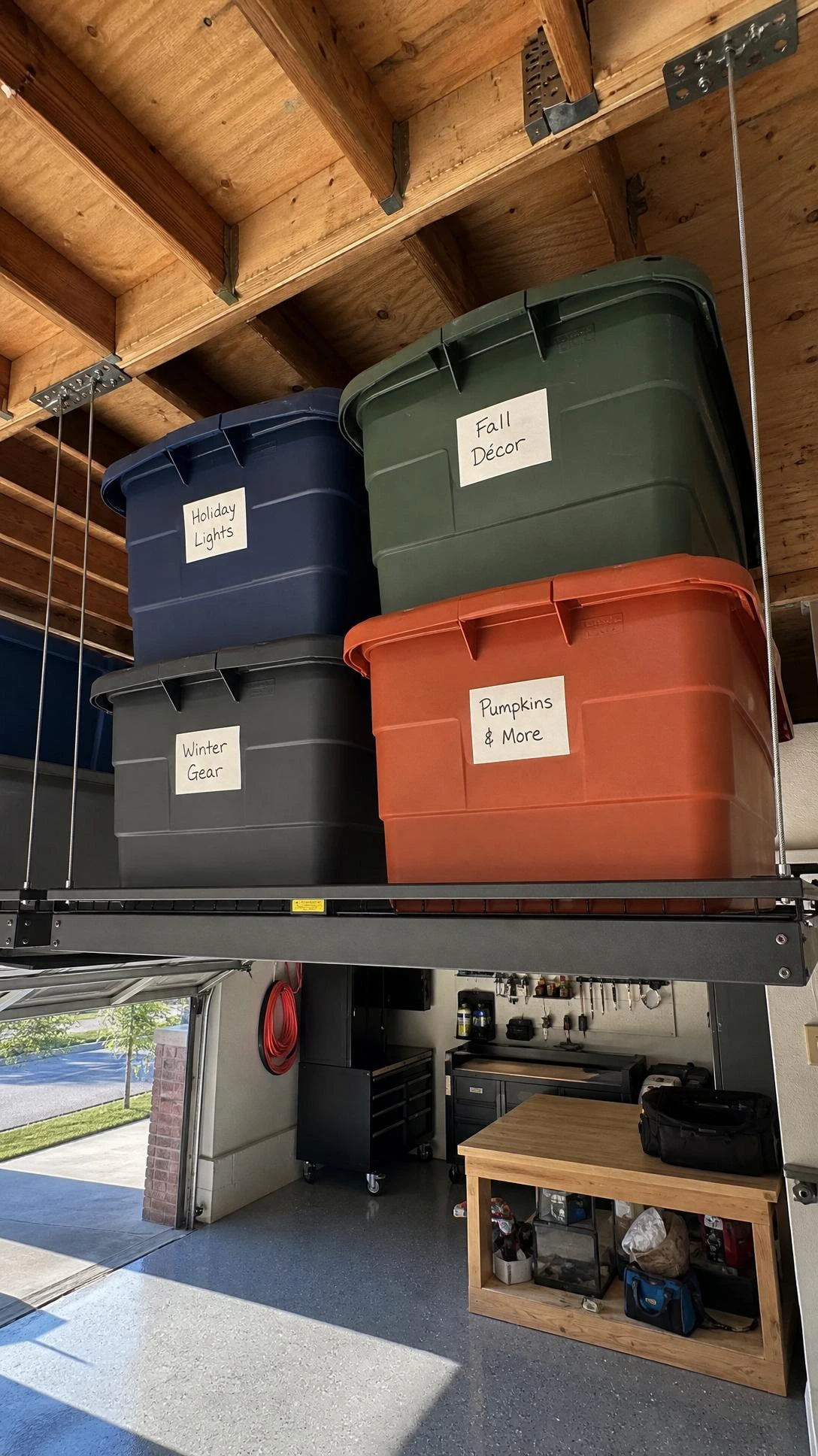Overhead garage ceiling storage platform with steel cable brackets holding large plastic seasonal bins stacked neatly — a beginner-friendly DIY that reclaims floor space