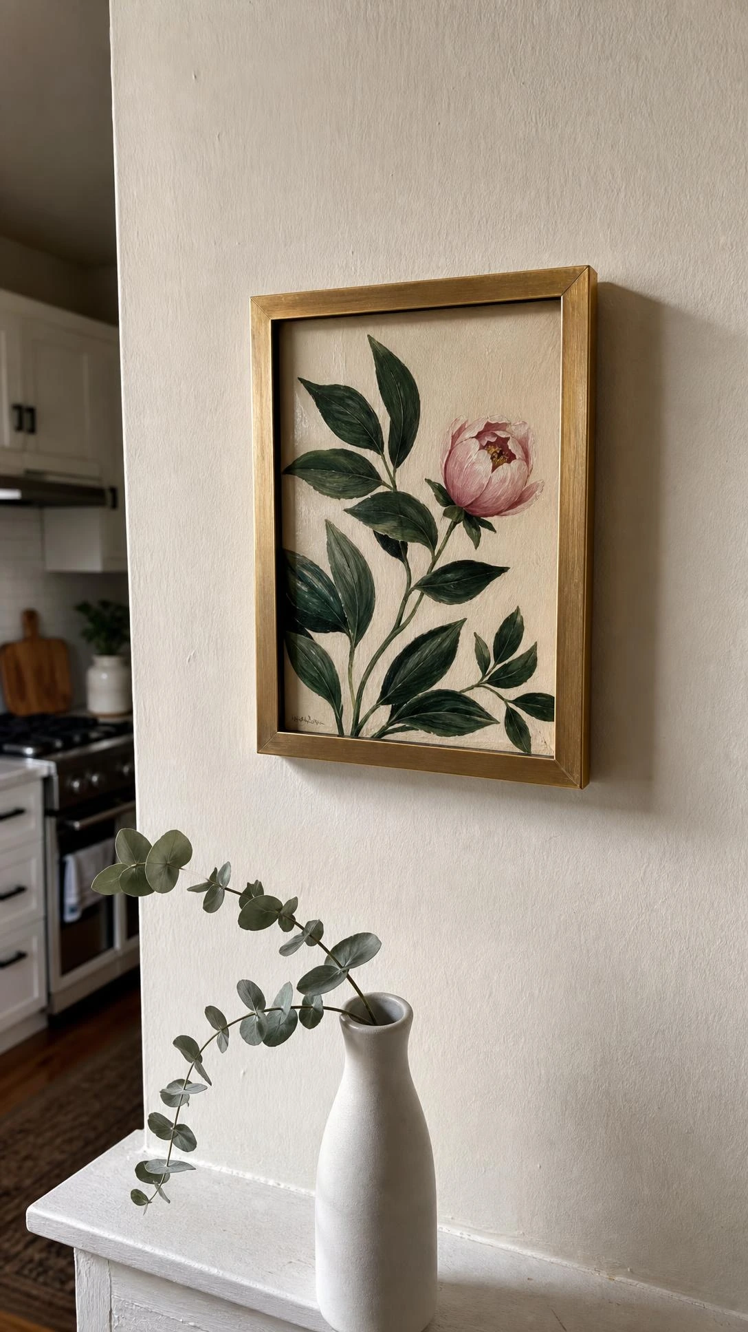Small brushed brass frame with botanical print — green leaves and blush bloom — hung slightly off-center on a white kitchen entry wall, proving one small piece of art can anchor an entire space