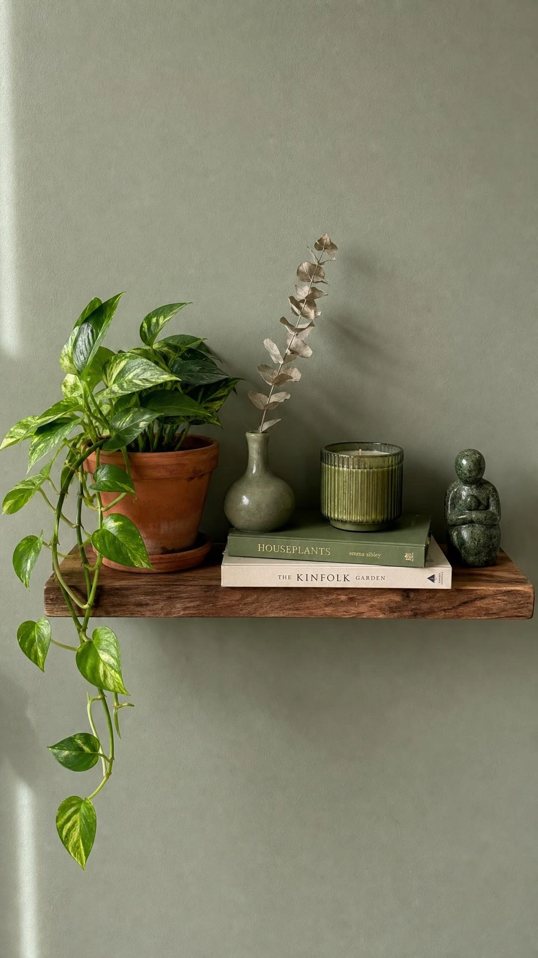 Small wooden floating shelf on sage wall styled with pothos, green ceramic vase, and olive candle for budget green aesthetic decor