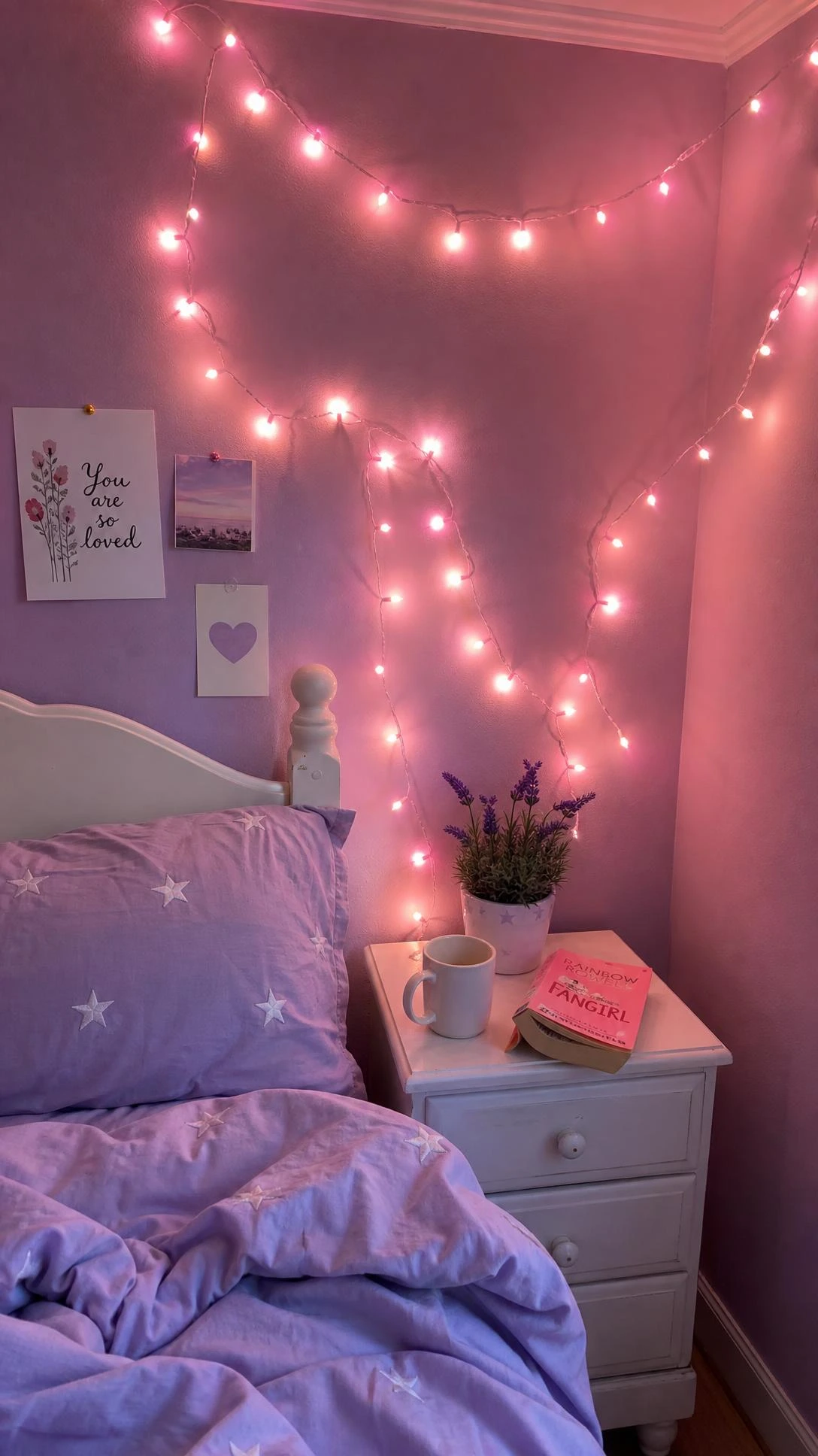 Soft pink string lights draped along a lavender wall above a white wooden bedside table, creating a warm glowing girls' bedroom corner on a budget