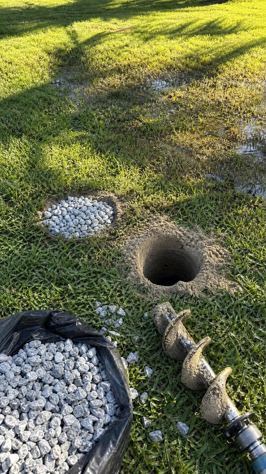 Freshly auger-drilled drainage hole in sandy Florida lawn soil with a nearby completed hole filled with angular gray gravel, solving standing water problems