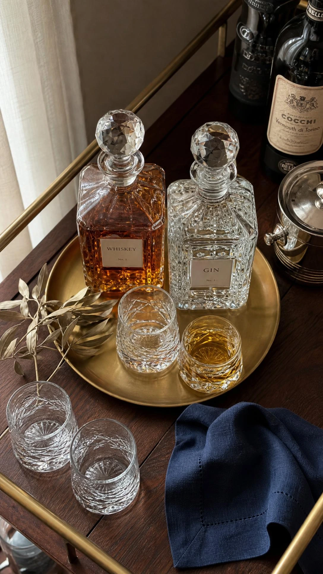 Overhead flat lay of a polished bar cart corner vignette with two faceted glass decanters holding amber whiskey and clear gin, styled in warm natural light