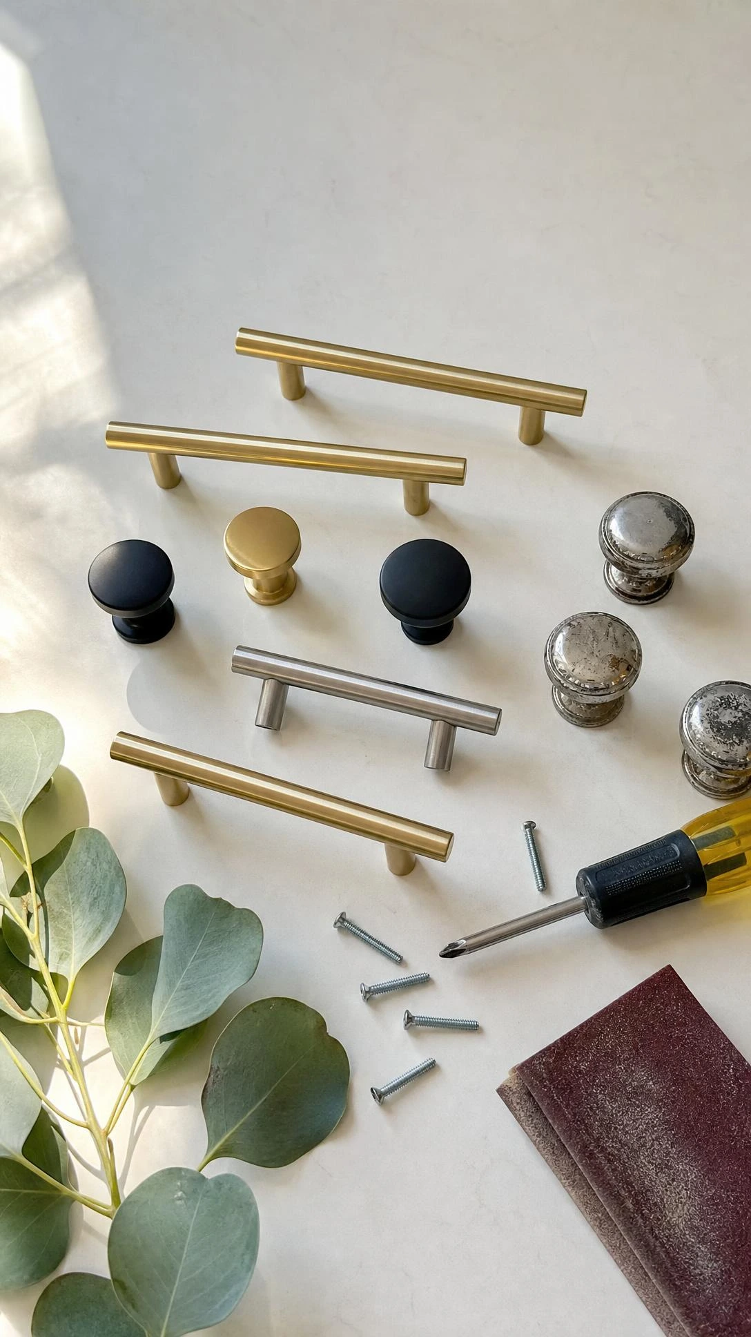 Flat lay of brushed brass pulls, matte black knobs, and a satin nickel bar pull on a white bathroom countertop showing affordable cabinet hardware upgrade options