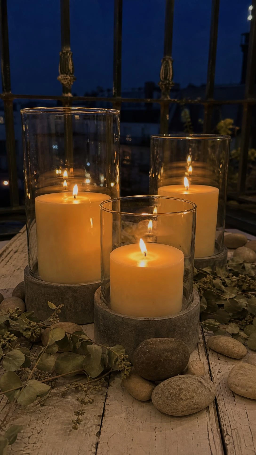 Three pillar candles in glass hurricane holders casting golden amber light on a weathered white balcony surface at dusk, a safe and elegant outdoor candle arrangement