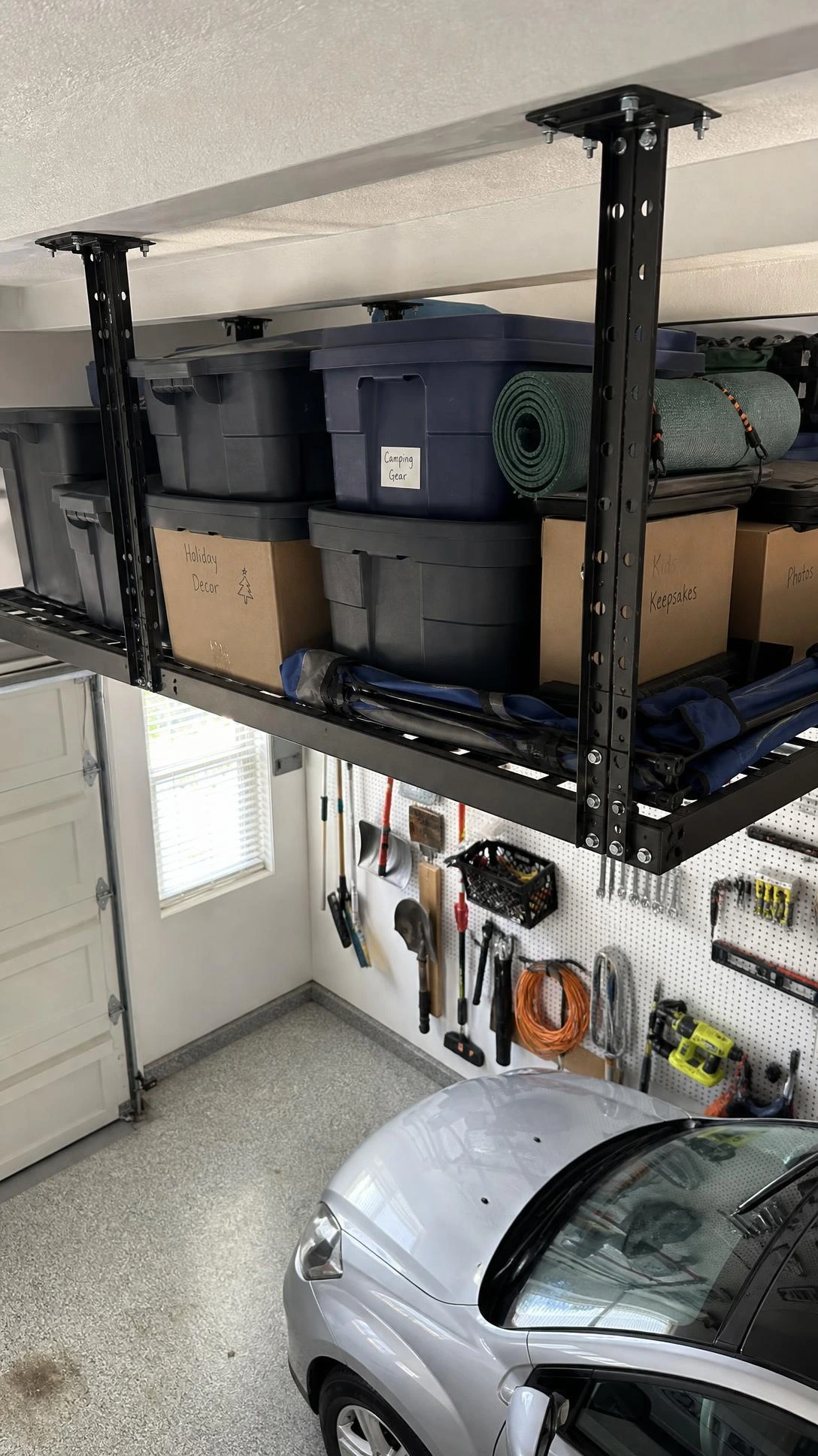 Heavy-duty black steel ceiling overhead storage rack mounted to white garage joists, holding organized gray bins and gear above the garage floor to reclaim every inch of vertical space