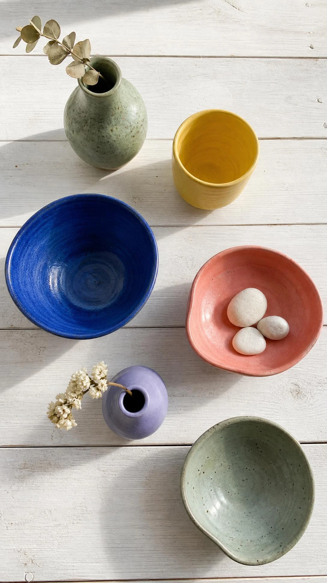 Overhead flat lay of handmade ceramics on a light oak shelf, featuring a cobalt blue matte bowl and sage green speckled stoneware arranged with minimalist intention