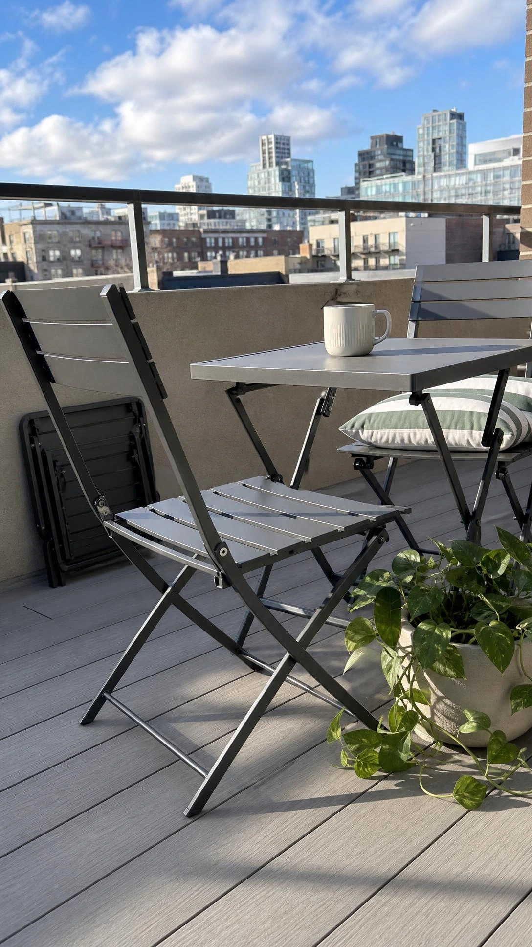 Slim slate gray foldable bistro table and two matching chairs fully open on a compact rooftop terrace, shot low-angle to show how foldable furniture maximizes tight outdoor spaces