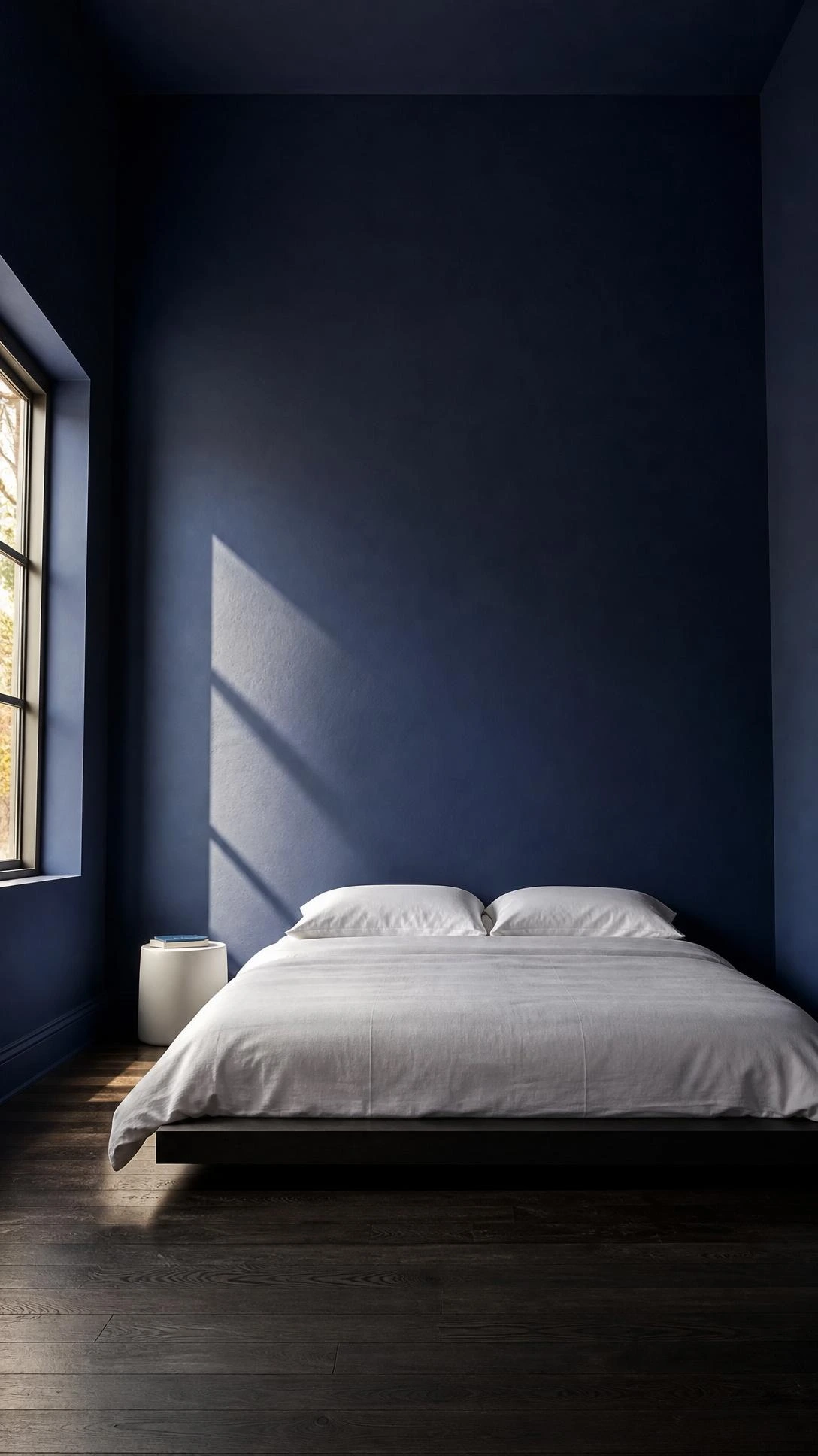 Deep navy blue bedroom with floor-to-ceiling blue walls, low platform bed, white linen duvet, and almost zero decor for a bold minimalist look