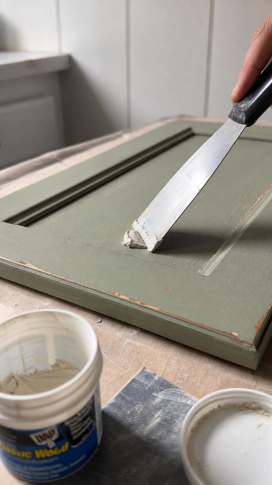 Putty knife pressing white wood filler into an old hardware hole on a sage green cabinet door, demonstrating the hole-filling step in a cabinet makeover