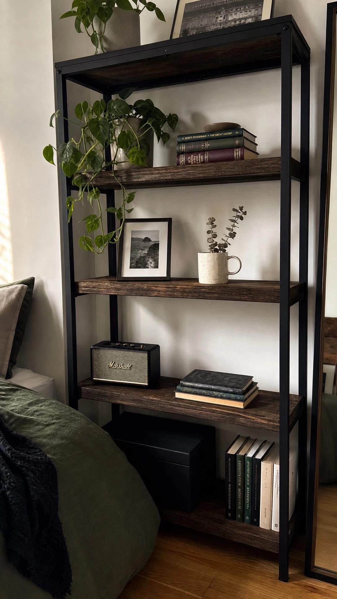 FJÄLLBO shelving unit styled with books and plants against a white bedroom wall, showing how raw wood and black steel add rustic texture without a renovation