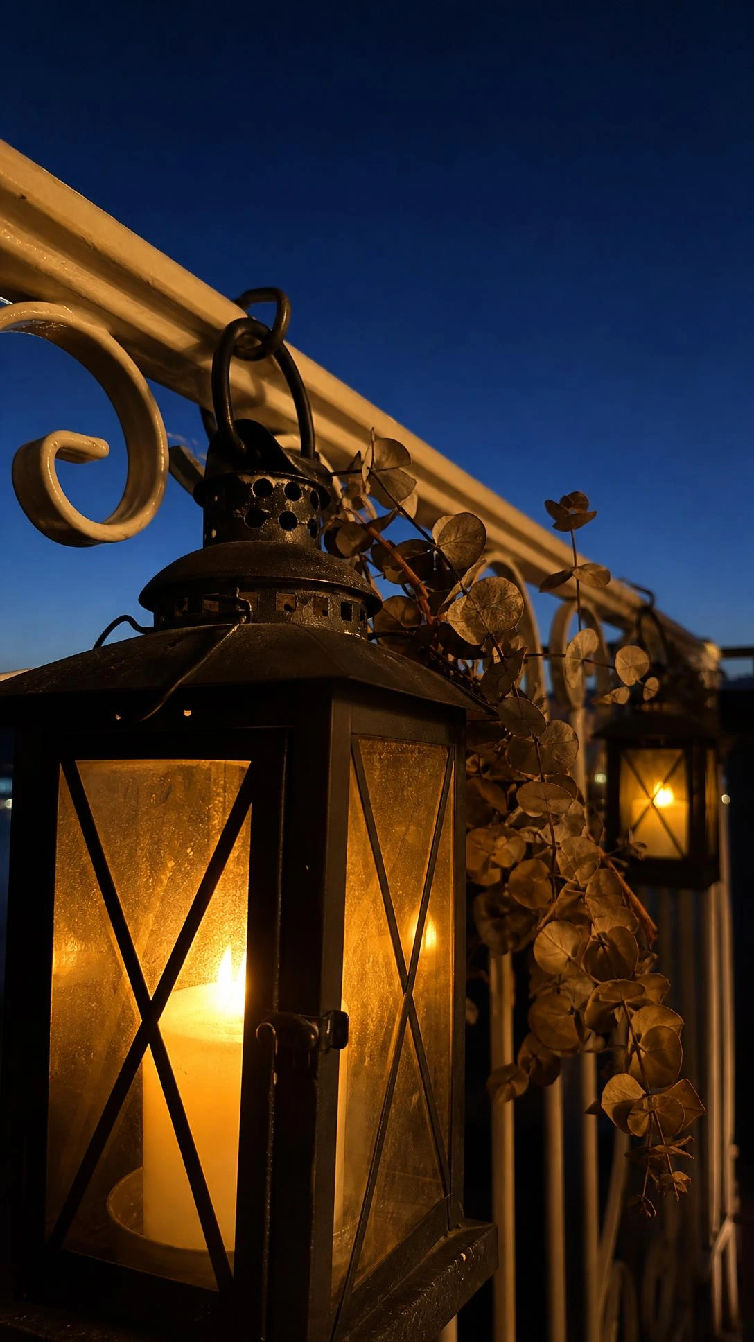 Vintage matte black iron lantern with clear glass panels hanging on a balcony railing at dusk, glowing with warm amber candlelight for cozy outdoor ambiance