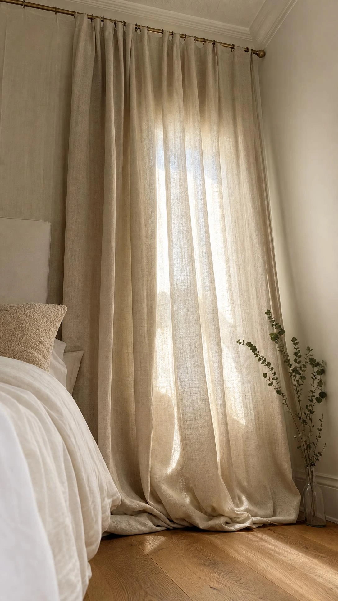 Floor-to-ceiling oat linen curtains pooling softly on hardwood floors in a serene white and beige bedroom, shot from low angle to emphasize their full draping height