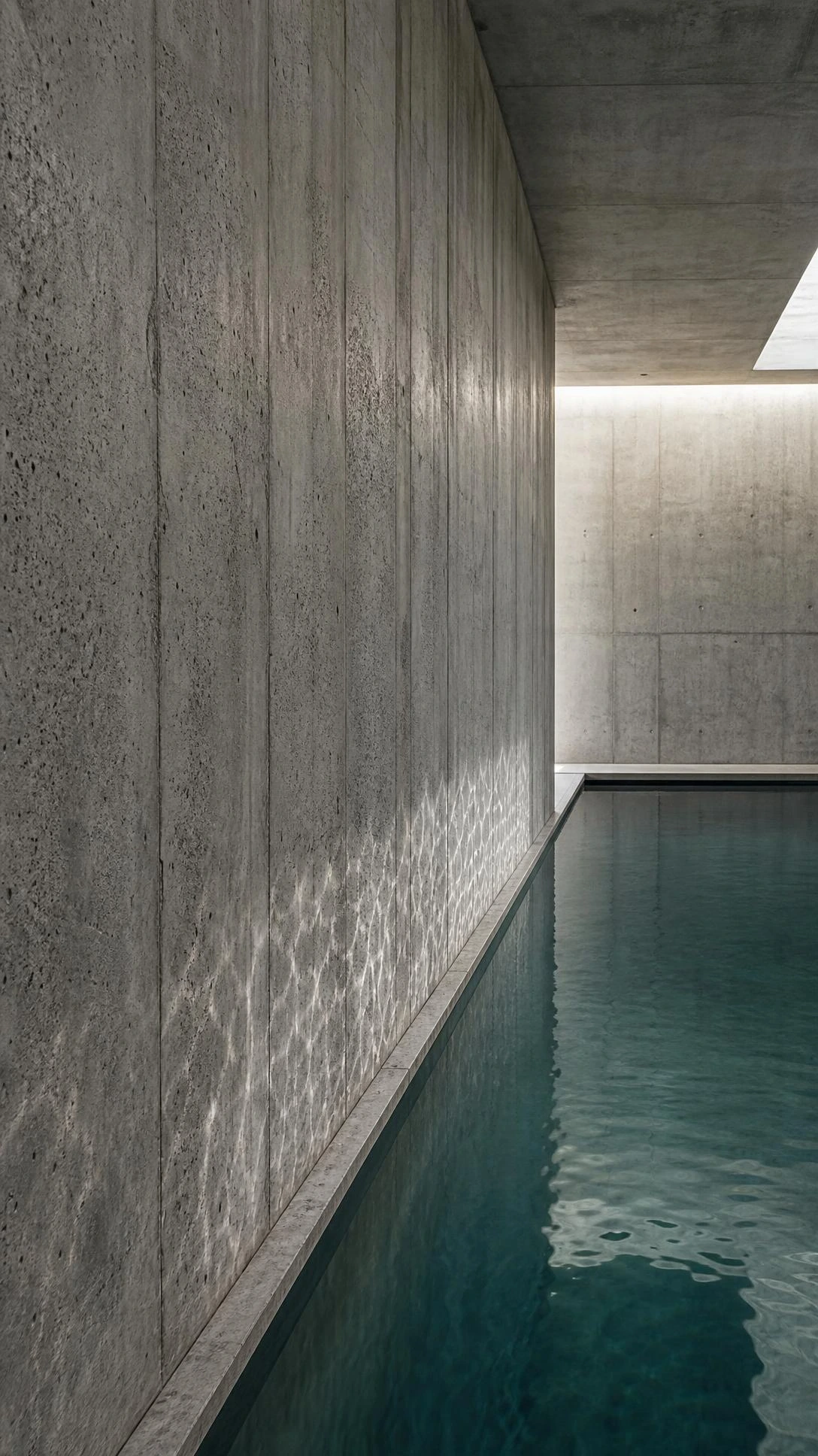 Raw concrete wall meets still water's edge in a minimalist indoor swimming pool, where natural skylight highlights the textured formwork lines and organic mineral tones