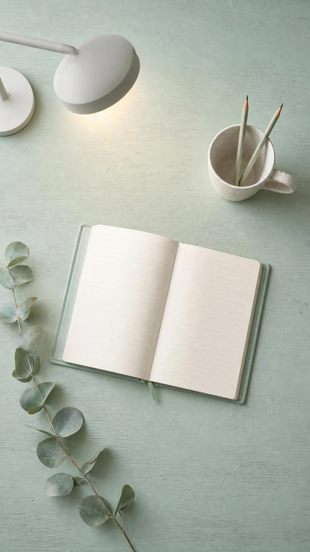 Flat lay of a small wooden desk painted in soft mint green holding a speckled ceramic mug, open notebook, and eucalyptus sprig — mint green paint brightening a compact home office