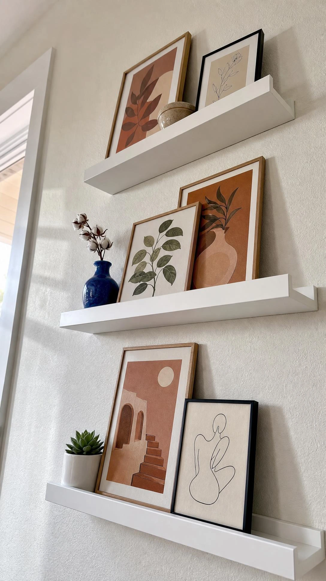Three white MOSSLANDA picture ledges mounted in a staggered arrangement on a painted wall, styled with leaning art prints in a bedroom gallery display