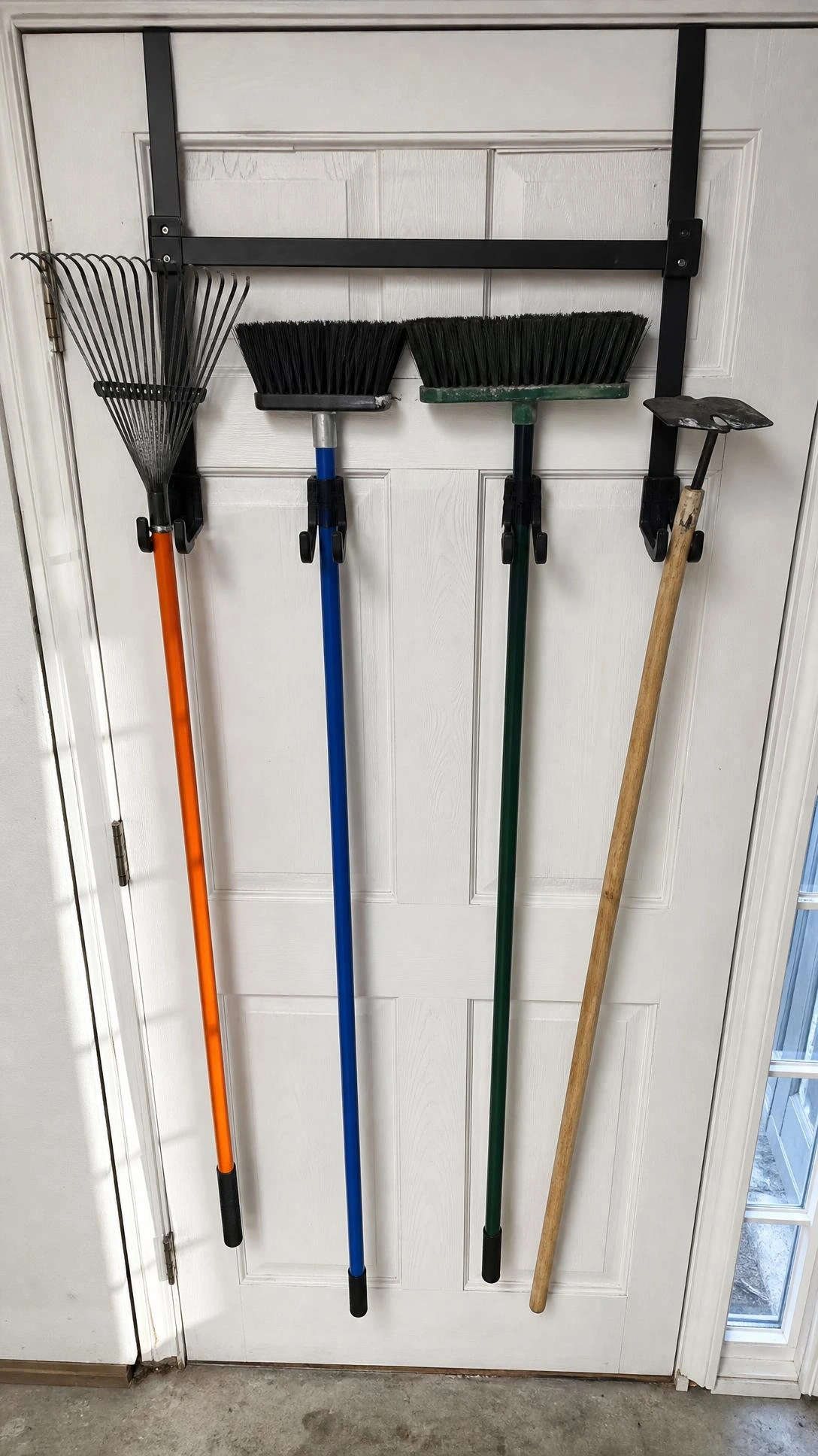 Over-door long-handled tool hooks on a white garage door holding a rake, flat-head shovel, and broom in a small garage corner, maximizing vertical storage