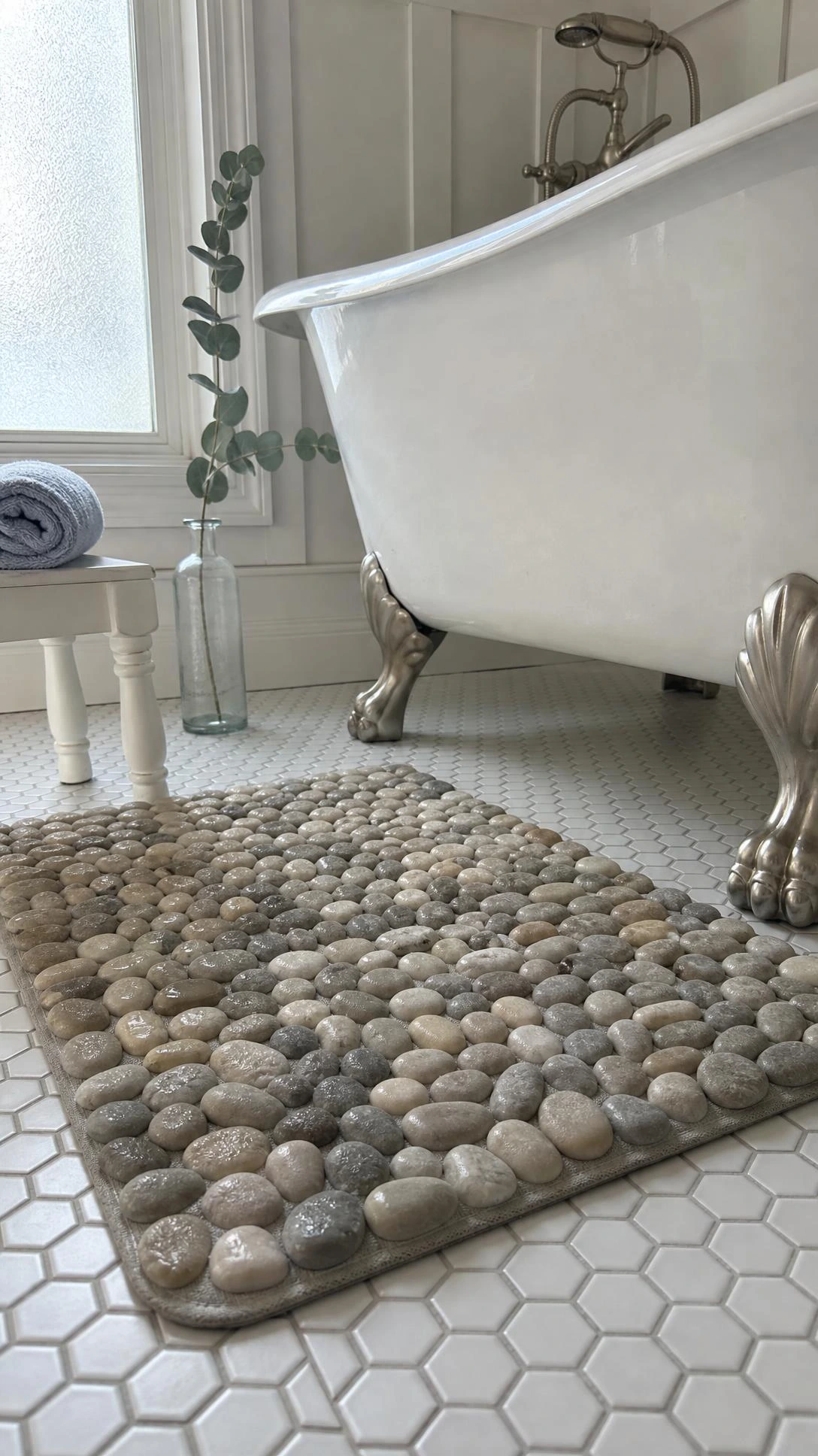 Pebble-textured bath mat in sandy taupe and warm gray tones placed in front of a white clawfoot tub, catching soft morning light for a coastal spa feel