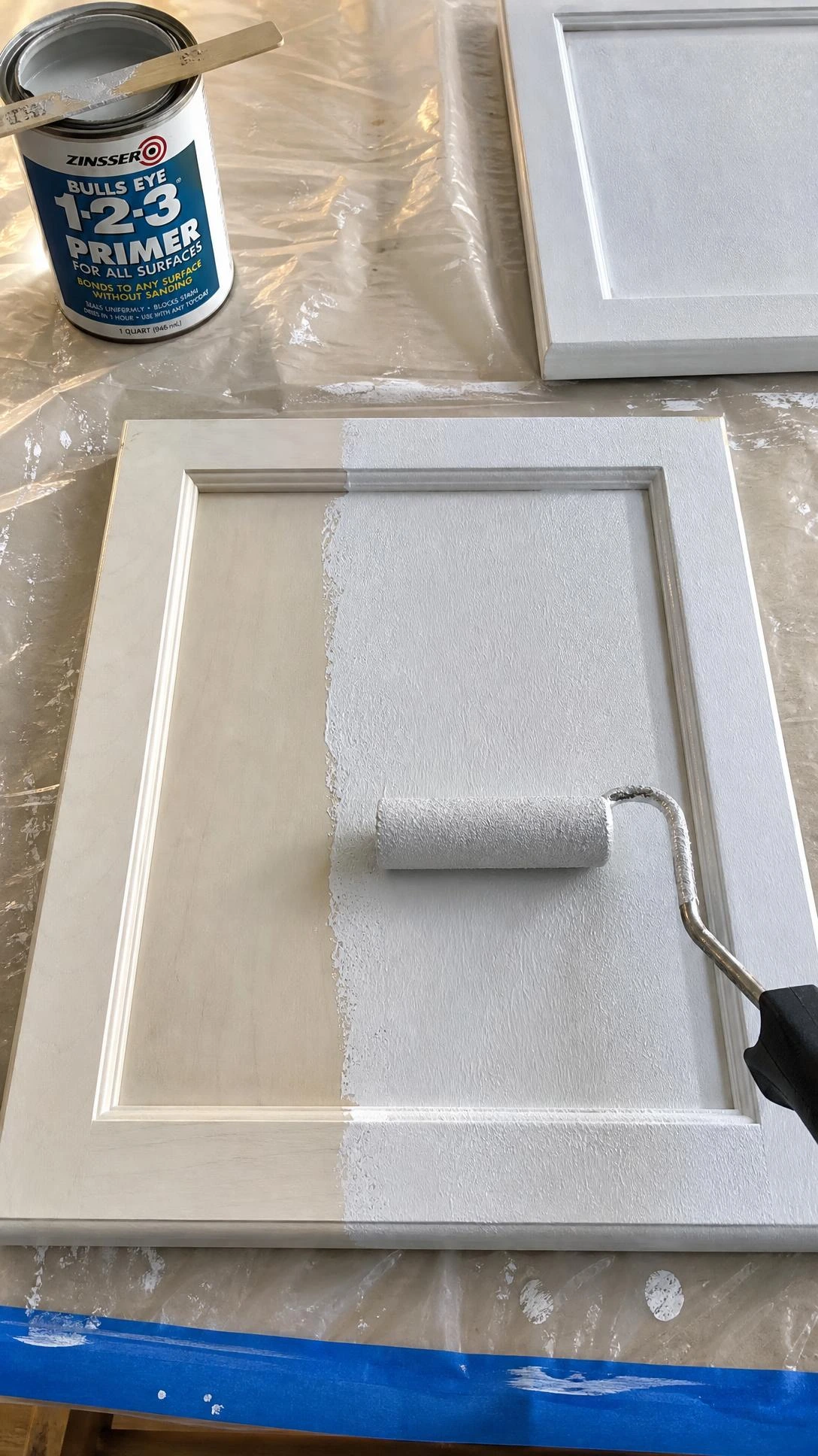Gray-white bonding primer being rolled onto a flat cabinet door, showing wet textured coverage on half the surface—essential prep step for a kitchen cabinet makeover