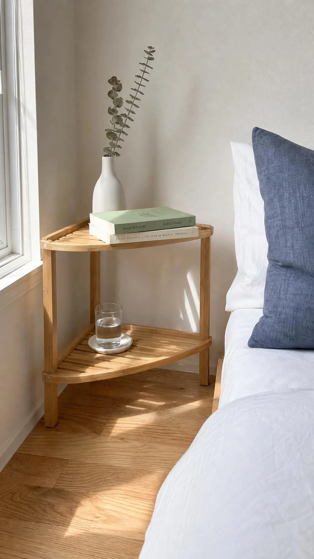 RÅGRUND corner bamboo table used as a minimalist bedside unit, tucked into a bedroom corner beside a low platform bed with white bedding