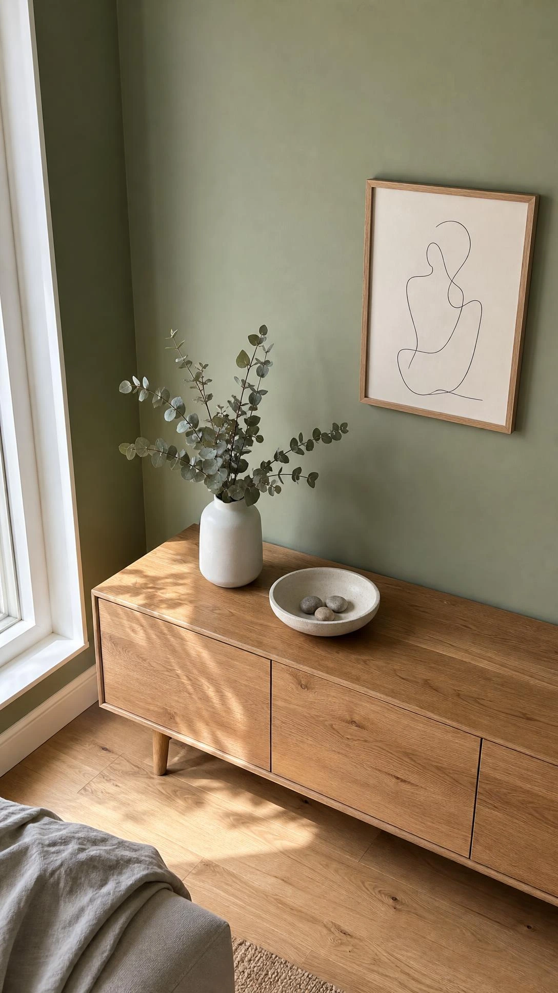 Sage green accent wall paired with warm wood furniture in a minimalist living room corner, showing how muted color adds calm without visual clutter
