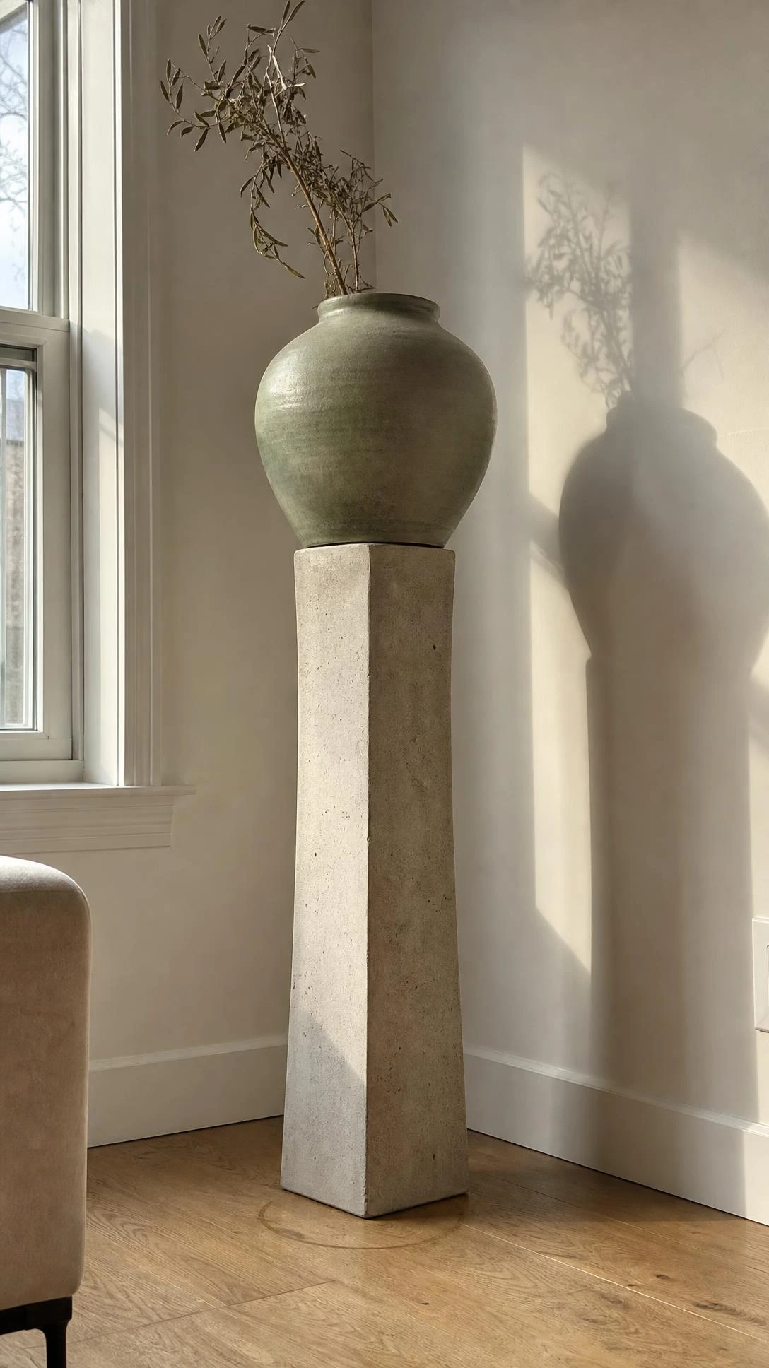 Tall concrete pedestal holding one oversized matte sage green ceramic vessel filling an empty living room corner with sculptural vertical impact