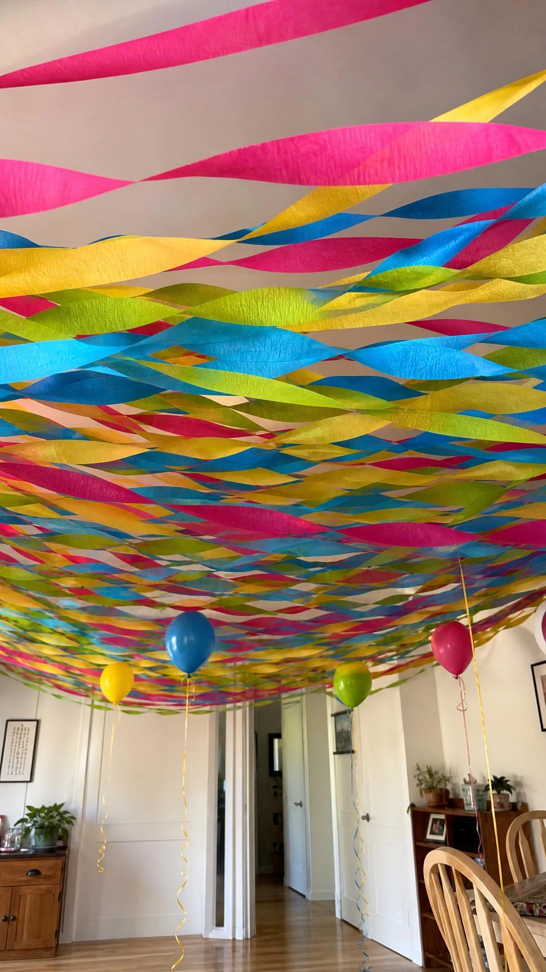 Colorful birthday party streamers in magenta, yellow, blue, and green crisscrossing a living room ceiling, seen from a child's eye-level view