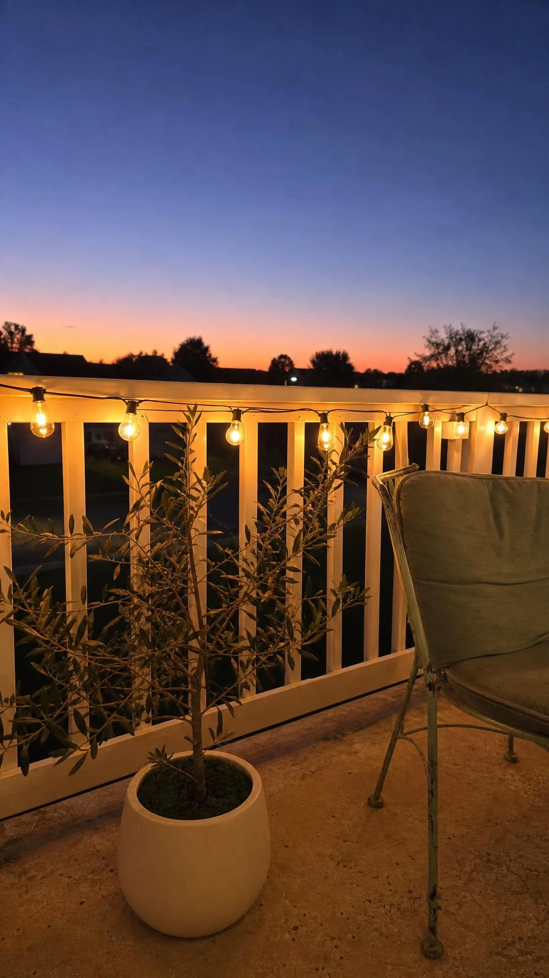 Warm Edison string lights draped along a white wooden balcony railing at golden hour, glowing amber-gold against a blue-purple evening sky