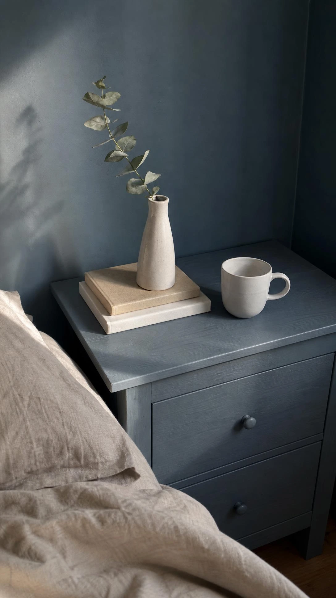 TARVA bedside table painted in dusty blue-gray blending into a matching wall, pine wood grain faintly visible beneath the matte finish