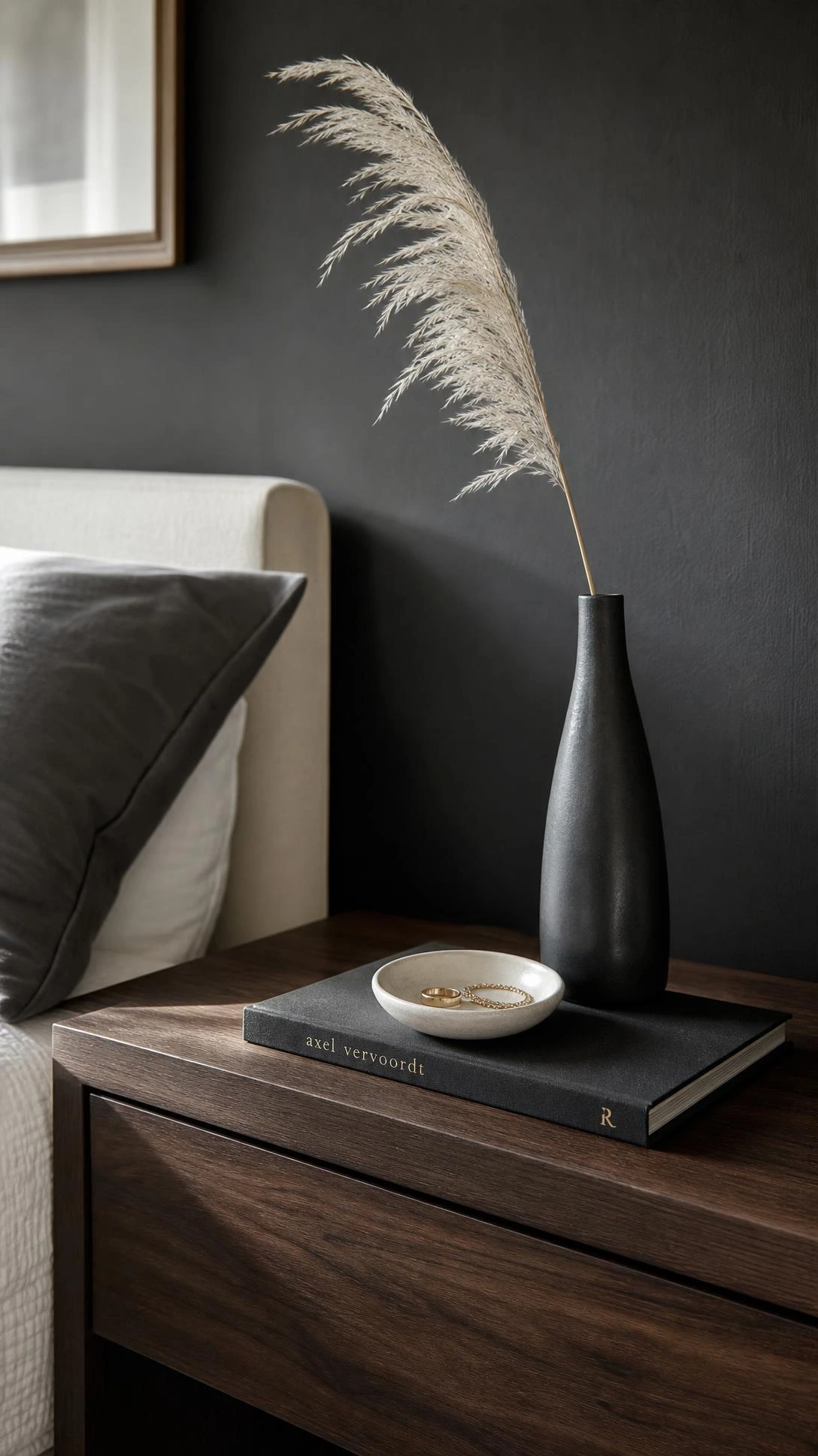 Modern luxury bedroom nightstand styled with matte black ceramic vase holding dried pampas, a porcelain trinket dish, and charcoal hardcover book — intentional minimalist bedside styling