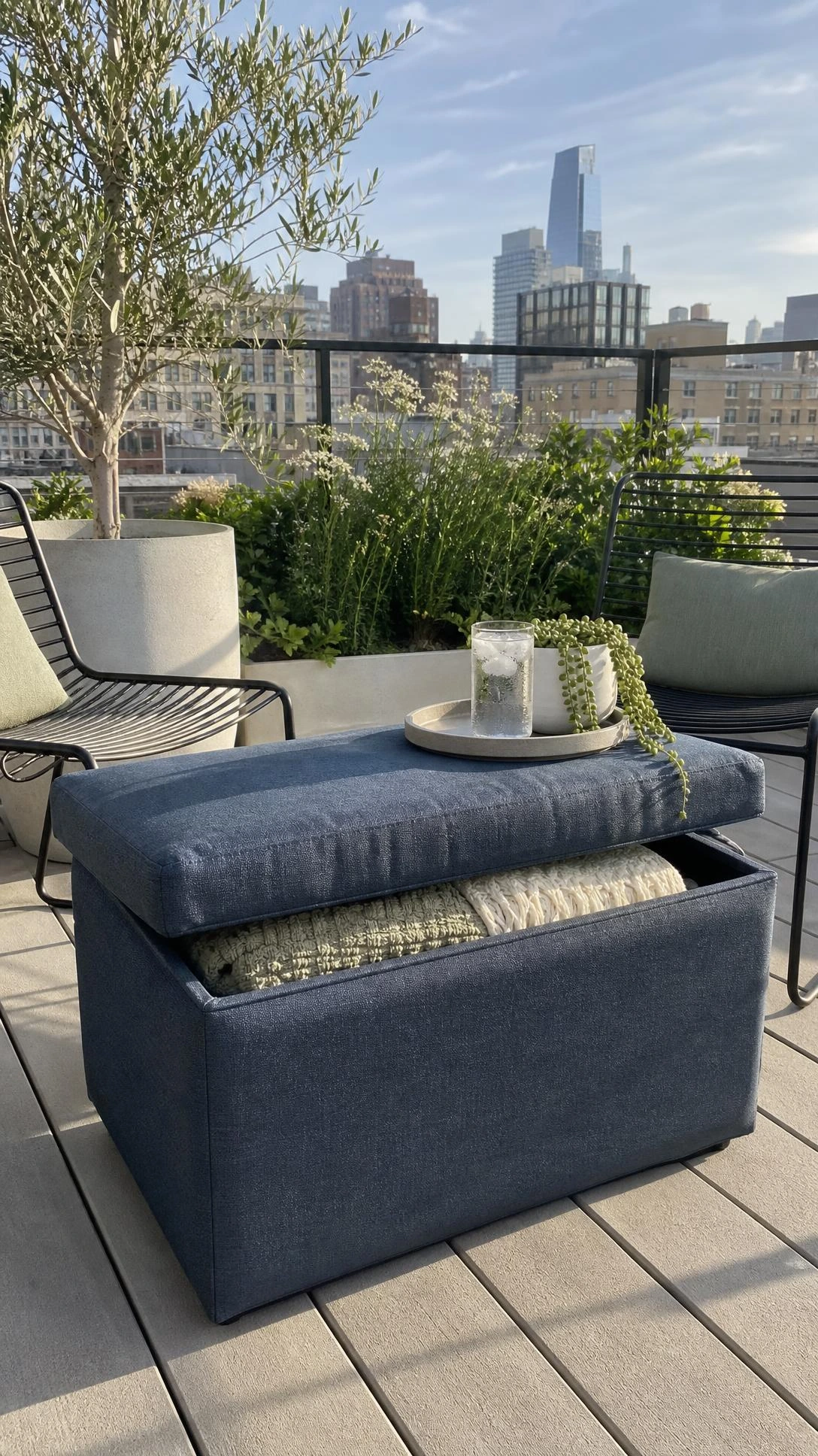 Weather-resistant slate blue Sunbrella storage ottoman on a small urban rooftop terrace, lid propped open to reveal hidden storage inside