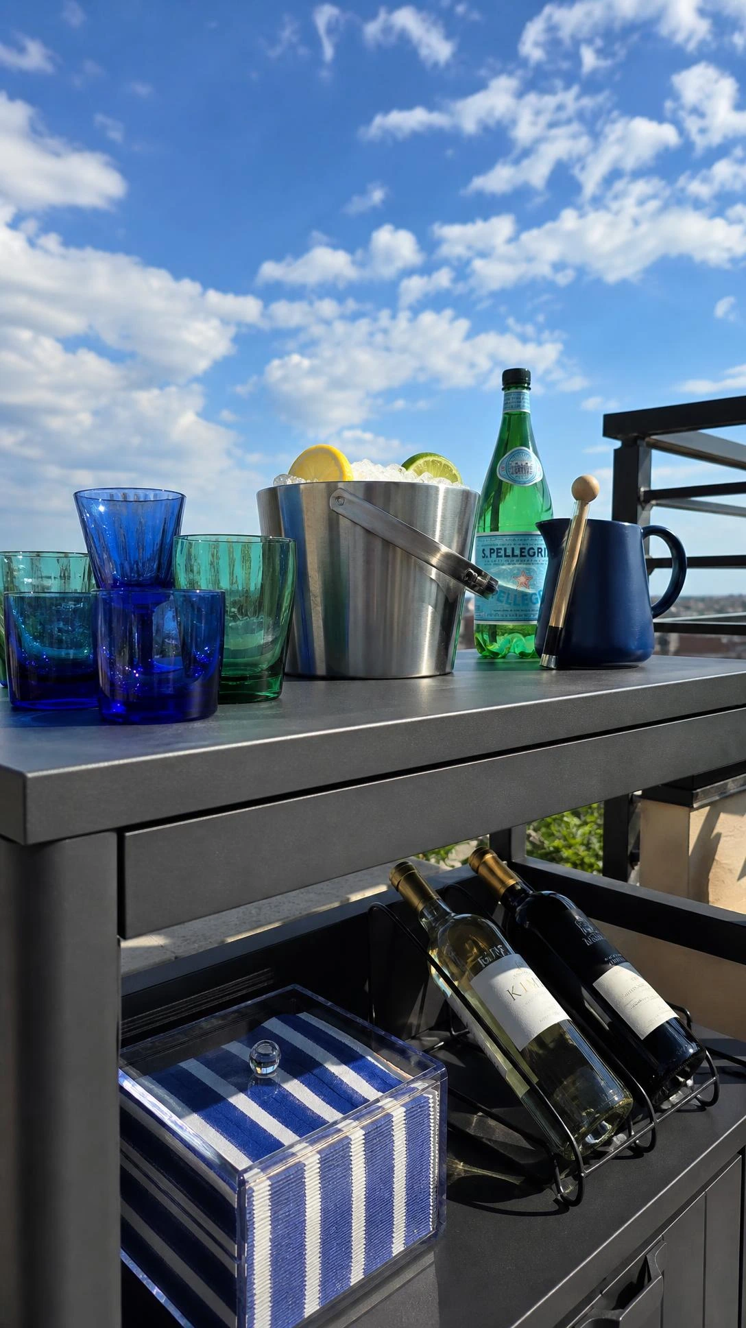 Low-angle view of a weatherproof rooftop drinks station with matte charcoal steel frame and slate-gray composite counter against a bright summer sky